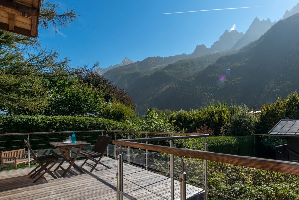 A spacious wooden terrace offers a panoramic view of the surrounding mountains, with a rustic table and folding chairs positioned for outdoor relaxation. Lush greenery and scattered trees enhance the tranquil setting, inviting moments of leisure under clear blue skies.