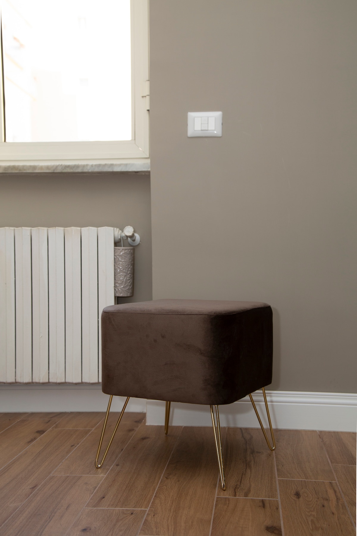 A compact brown upholstered stool with gold legs is positioned beside a window. The wall behind is a soft, neutral tone, and a radiator is visible in the background, enhancing functionality in the space.