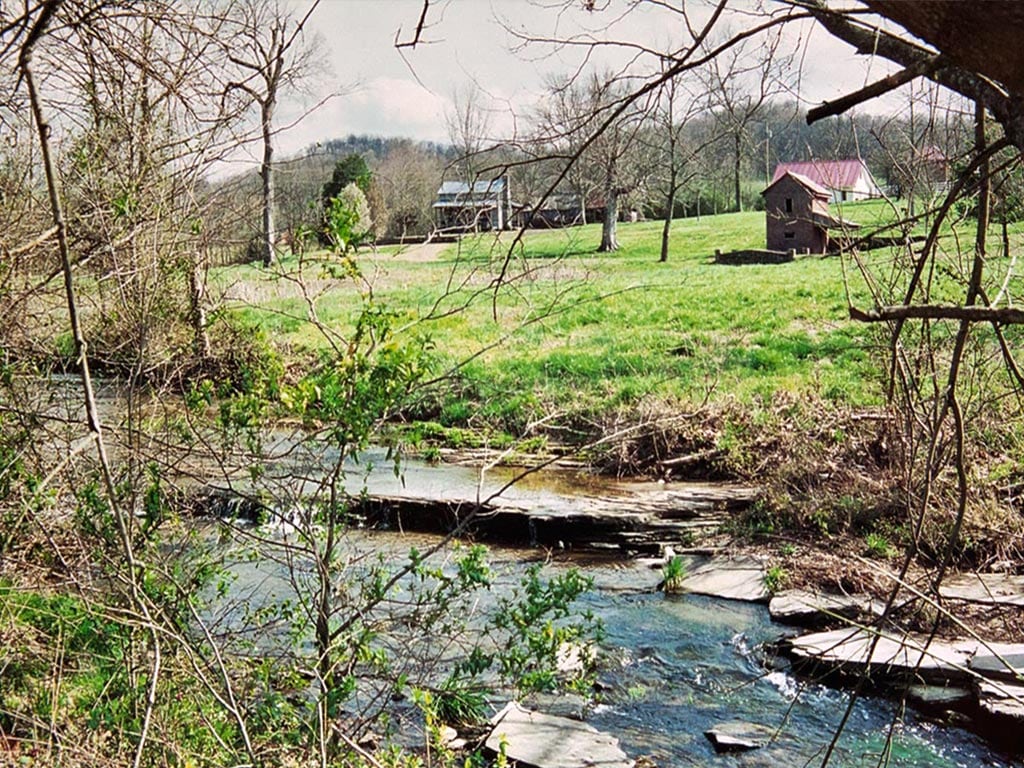 A serene creek runs through the foreground, gently flowing over smooth stones. In the background, a spacious green field is visible, with a cabin and outbuilding nestled among trees. The scene is framed by bare branches, hinting at a peaceful rural landscape.