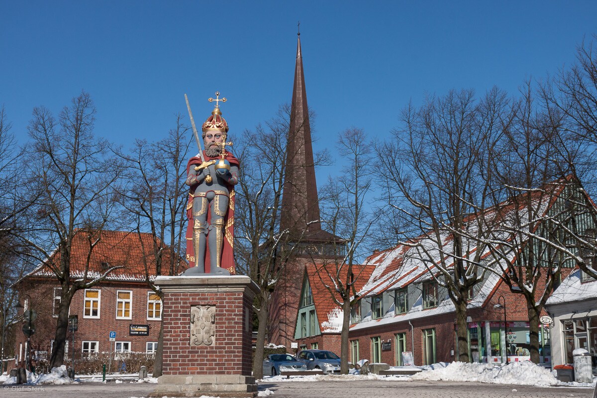 A statue of a knight stands prominently, adorned in regal attire and holding a sword. Behind it, a tall church spire reaches towards the clear blue sky, while red-roofed buildings and bare trees surround the area, indicating a winter season.