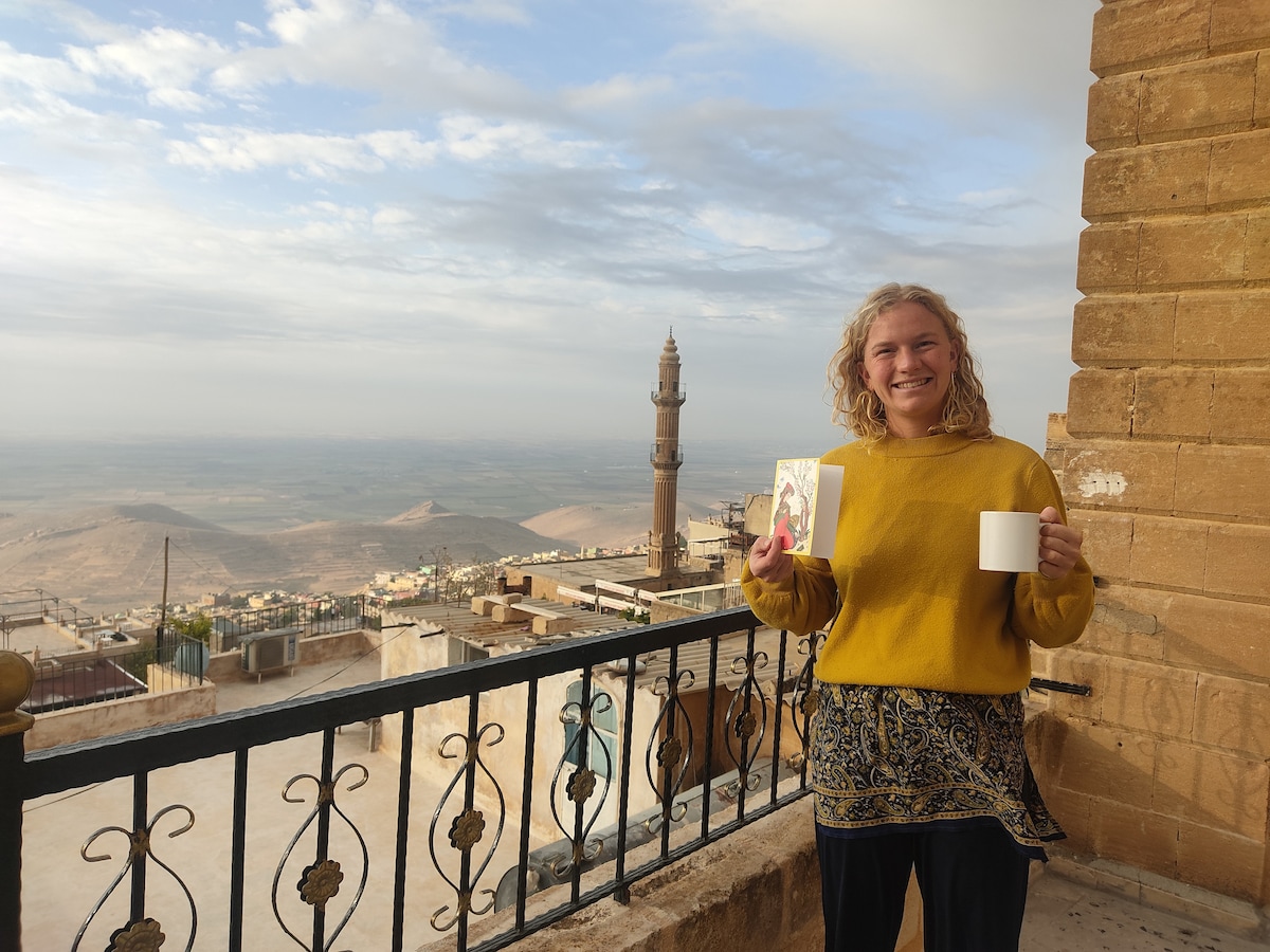 A person enjoys a scenic view from a terrace, holding a beverage and a card. The landscape features historical architecture and rolling hills extending towards the horizon, with a prominent minaret visible in the background.