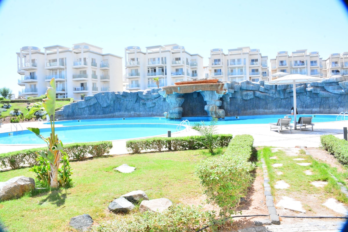 A large outdoor pool is surrounded by lush greenery and well-maintained landscaping. Comfortable lounge chairs are positioned under an umbrella beside the water. The backdrop features modern multi-story buildings and a decorative rocky structure, blending with the serene environment.