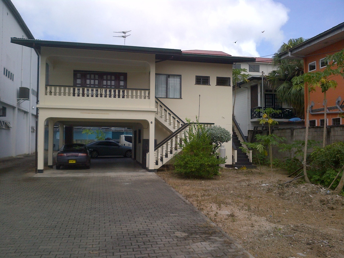 A two-story building is featured with a covered entrance and a staircase leading to the upper level. A parking area is visible beneath the structure, accompanied by greenery and a gravel pathway. The surrounding buildings and sky complete the view.