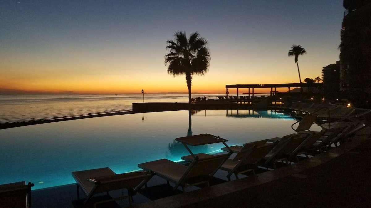 The image captures a serene evening view featuring a tranquil infinity pool reflecting the vibrant colors of sunset. Palm trees frame the scene, and sun loungers are positioned nearby, inviting relaxation by the water's edge. The ocean stretches beyond with gentle waves.
