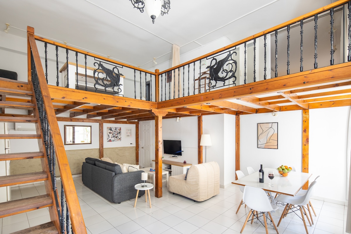 An open living area showcases wooden beams and a staircase leading to a lofted space. Sofas are arranged around a coffee table, while a dining table with chairs is positioned nearby. A television is mounted on the wall, and decor includes framed artwork.