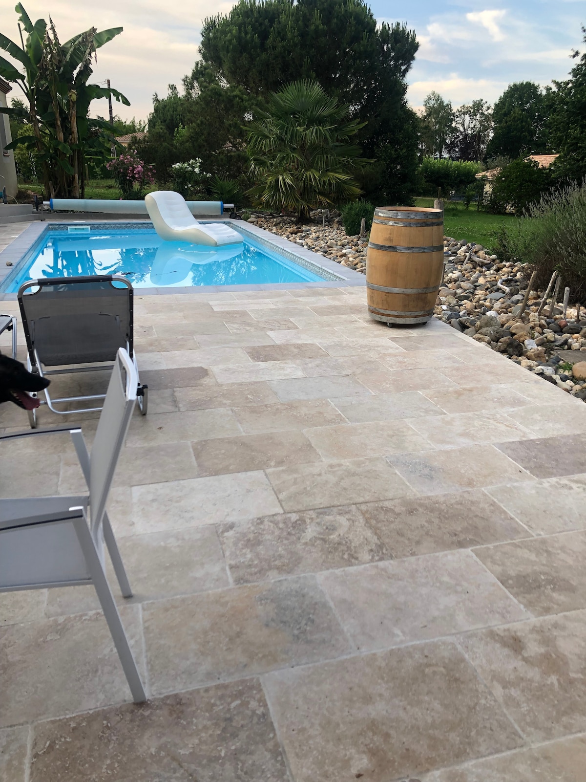 A swimming pool is set amid a landscaped area, surrounded by stone features and greenery. A lounge chair is positioned beside the pool on a tiled patio. A wooden barrel adds a rustic touch to the outdoor space, enhancing the serene environment.