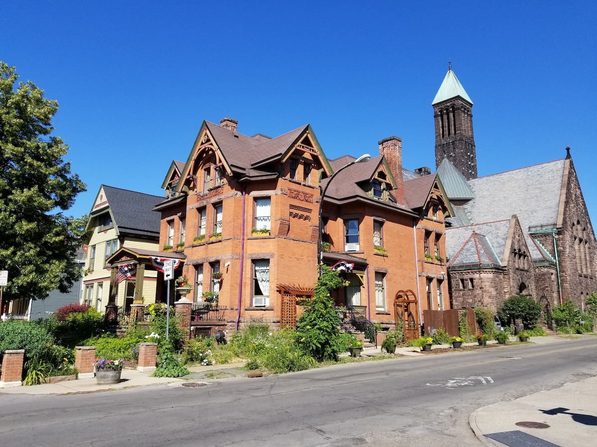An inviting view of a historic street lined with distinct architectural styles, showcasing a rehabilitation of an 1885 Queen Anne home with ornate brackets and large restored windows. Lush greenery and flower planters add to the vibrant streetscape. A church tower is visible in the background.