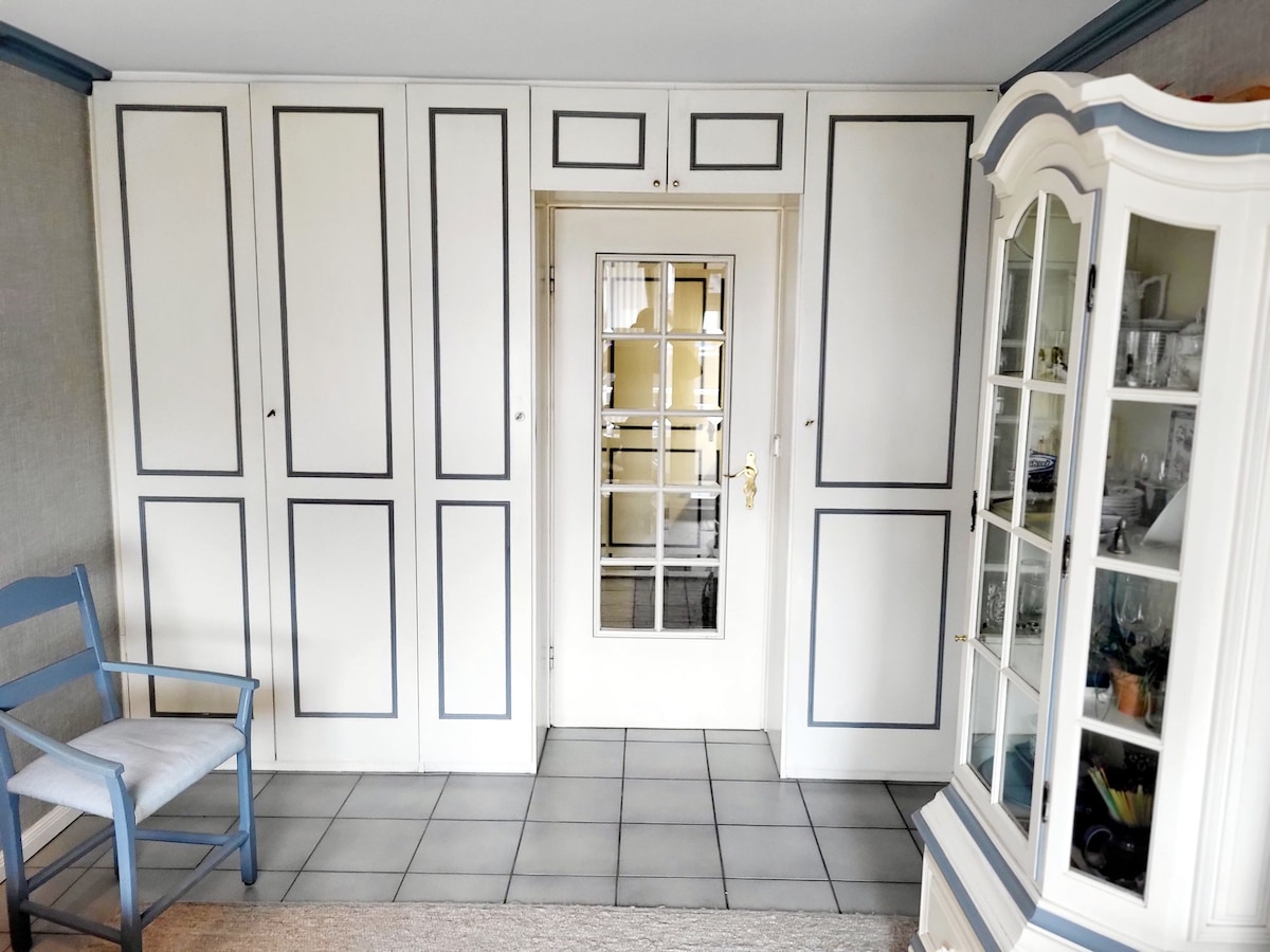 A bright entrance area is highlighted by a mirrored door leading to the interior. Built-in cabinets with white and grey accents offer ample storage. An elegant glass-front cabinet displays a variety of items. A light blue chair is positioned next to the cabinets on the tiled floor.