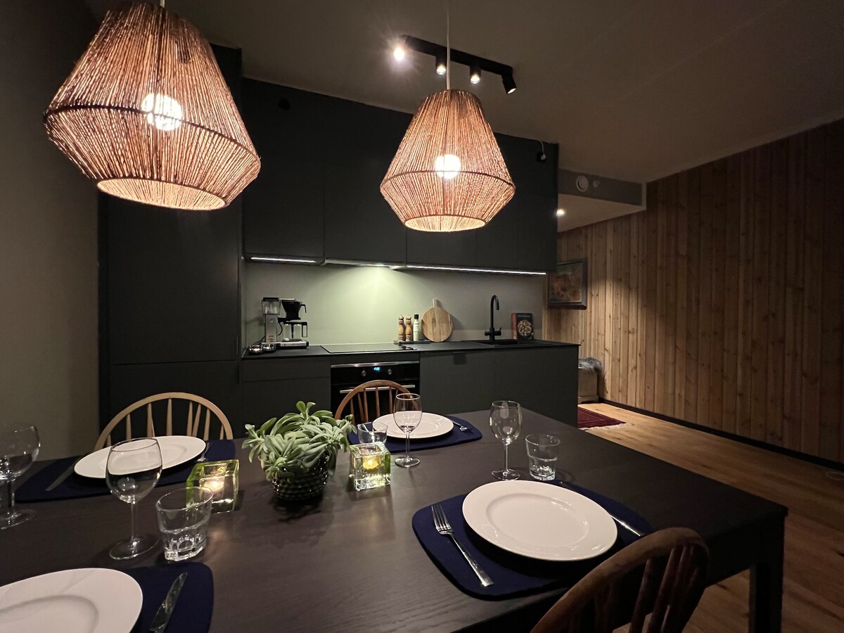 A modern kitchen is displayed with a dark cabinetry, featuring three woven pendant lights above a dining table. The table is set with white plates and glassware, complemented by small plants and candles as centerpieces. Warm wood accents adorn the walls of the inviting space.