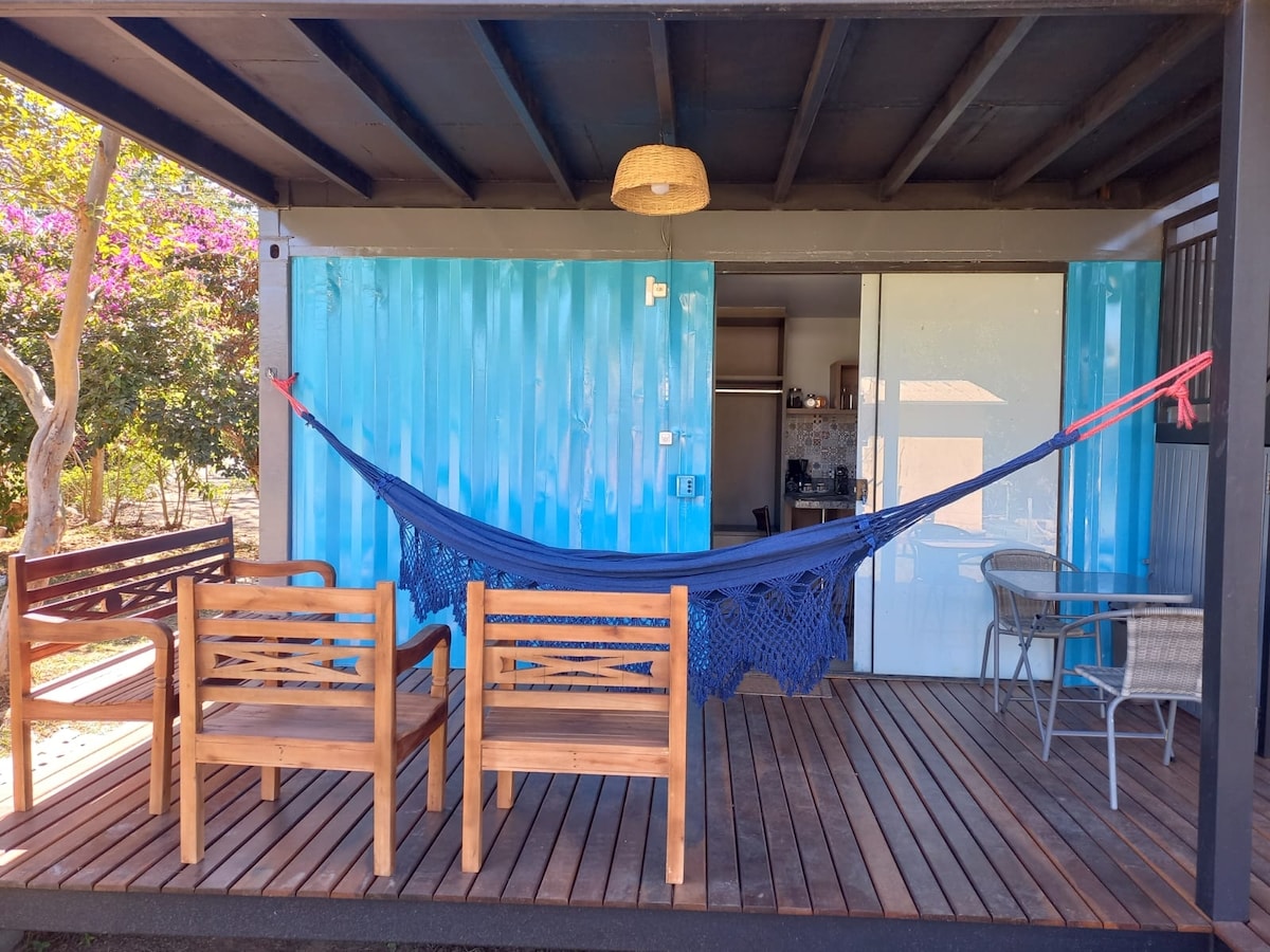 A covered porch area features a blue hammock suspended between two posts, complemented by several wooden seating options. The backdrop includes brightly painted walls and a glimpse of interior space, enhancing the area’s relaxed ambiance.