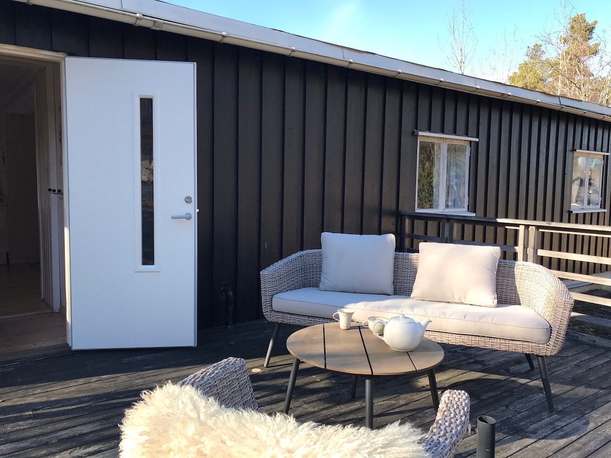 An outdoor seating area is visible on a wooden deck, featuring a comfortable sofa and a round table. A soft rug is placed beneath the table, and a door leading inside is present on the left. Natural light highlights the inviting and peaceful surroundings.