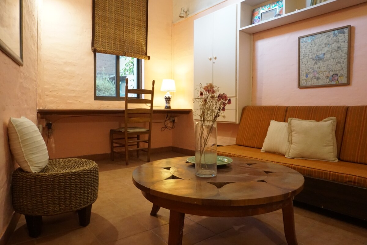 A charming living area includes a comfortable sofa dressed in earthy tones and a round wooden coffee table. A simple desk with a chair is positioned near the window, providing a view of the outside. Soft lighting is offered by a lamp placed on a side table.