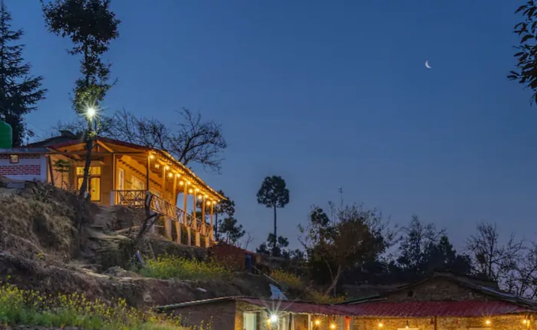 A charming hillside property is illuminated by soft lighting, set against a twilight sky with a crescent moon. The structure features a wraparound porch, with trees and hills in the background, creating a serene atmosphere surrounded by nature.
