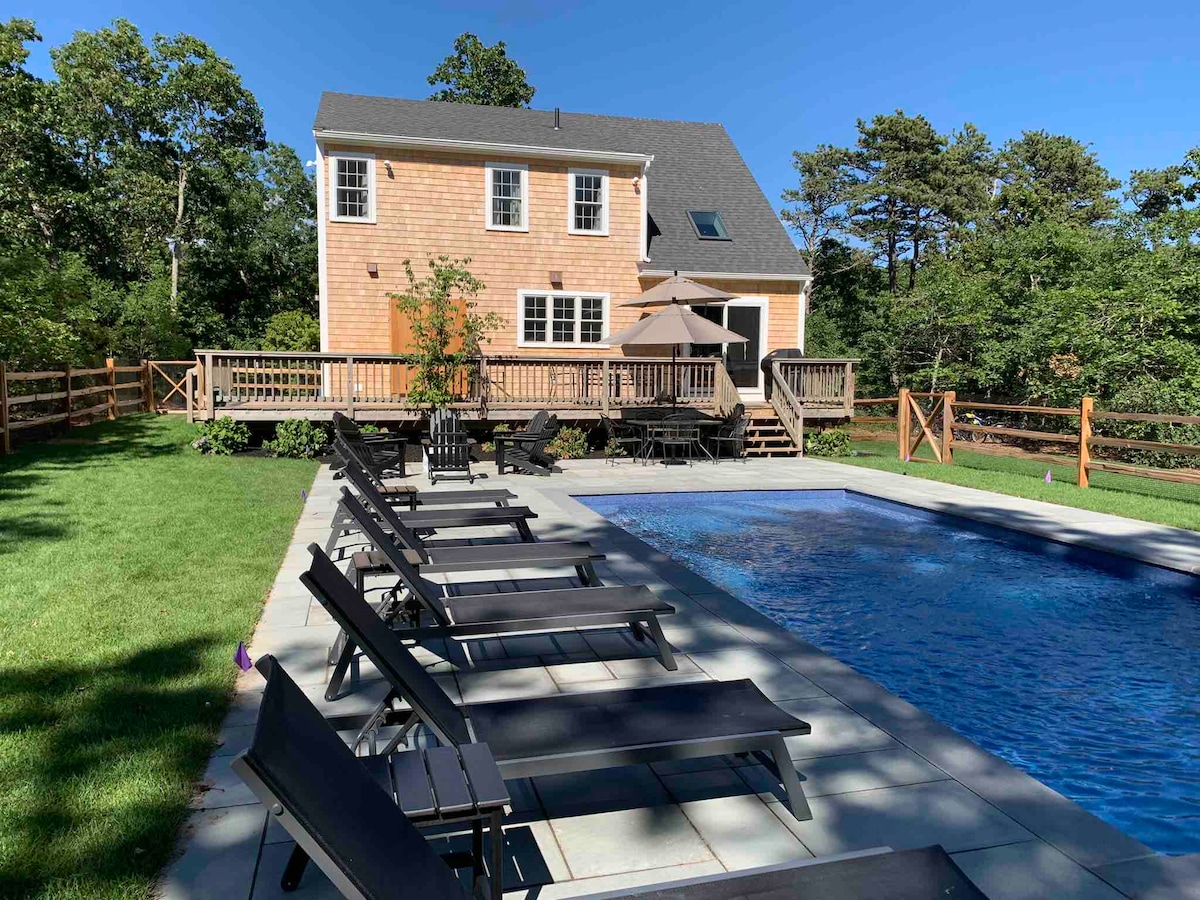 A colonial saltbox style home is shown with light wood siding, complemented by multiple windows. A deck is visible on the side, leading to a swimming pool surrounded by lounge chairs. Lush greenery frames the property, enhancing the outdoor space.