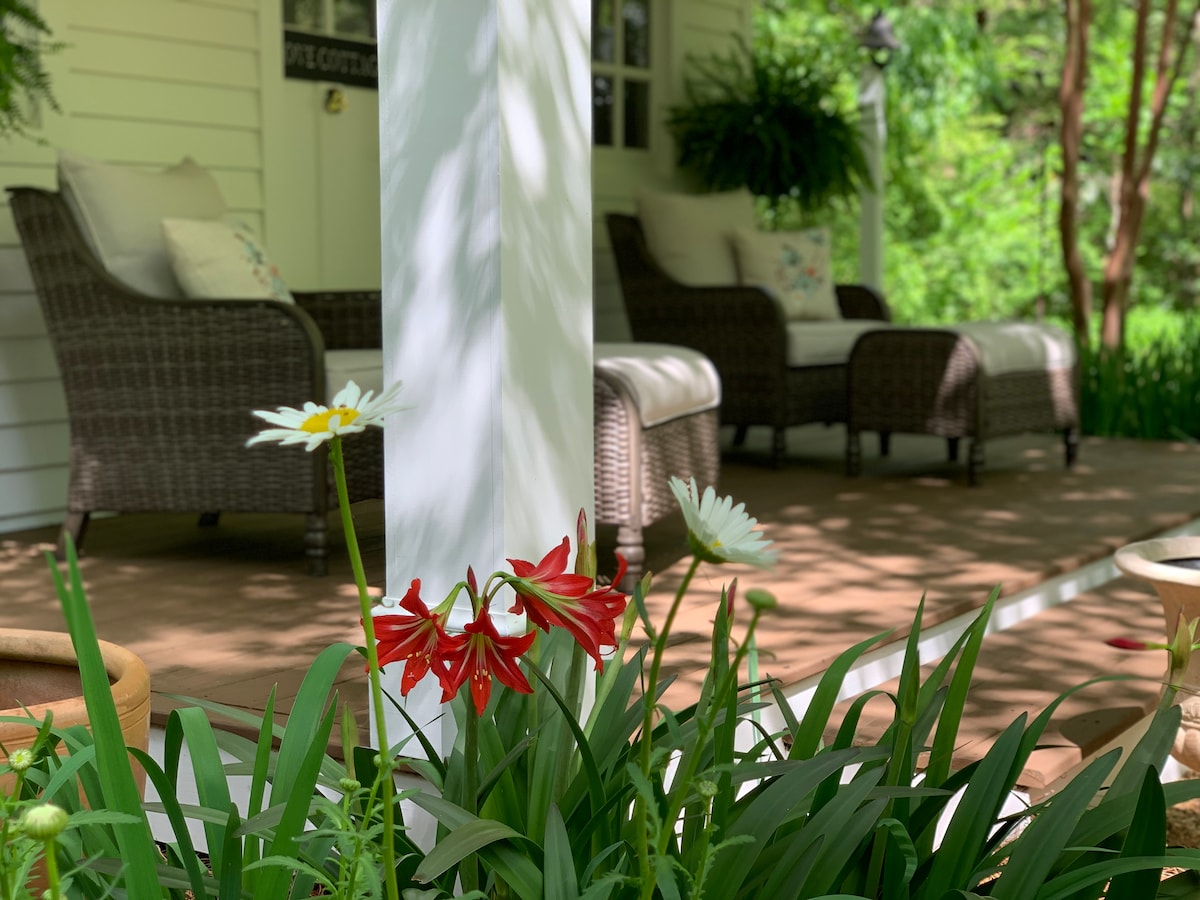 A welcoming front porch features two comfortable wicker chairs with soft cushions, positioned beside lush greenery and blooming flowers. Natural light filters through the surroundings, highlighting the inviting atmosphere for relaxation and enjoyment.