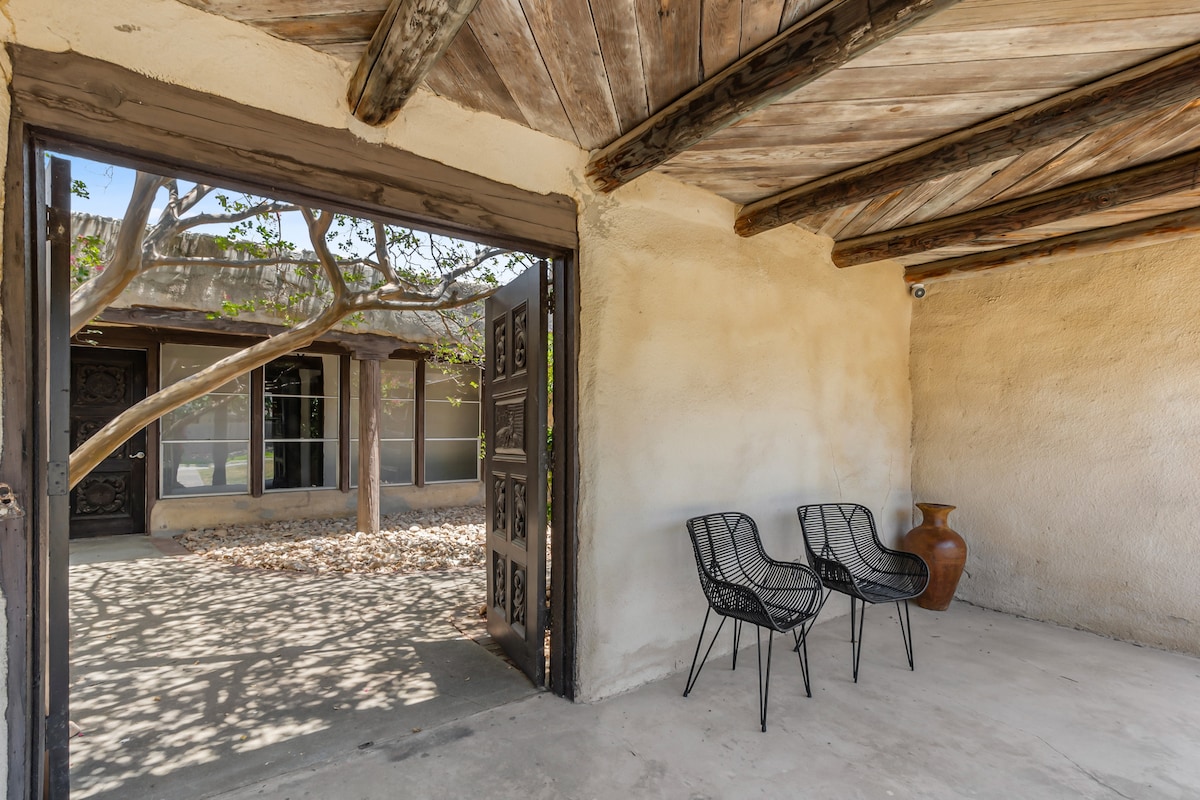 An entrance area features two black wire chairs positioned beside a clay pot. The rustic adobe walls and wooden ceilings complement the natural light streaming through the door, enhancing the connection with the tranquil courtyard view.