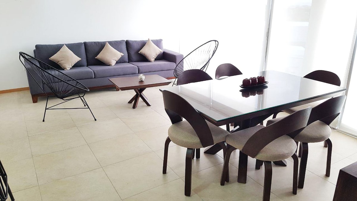 A spacious living area features a gray sofa with decorative cushions, paired with a glass dining table surrounded by six upholstered chairs. Two black, woven lounge chairs complement the modern aesthetic, with natural light filtering through large windows.