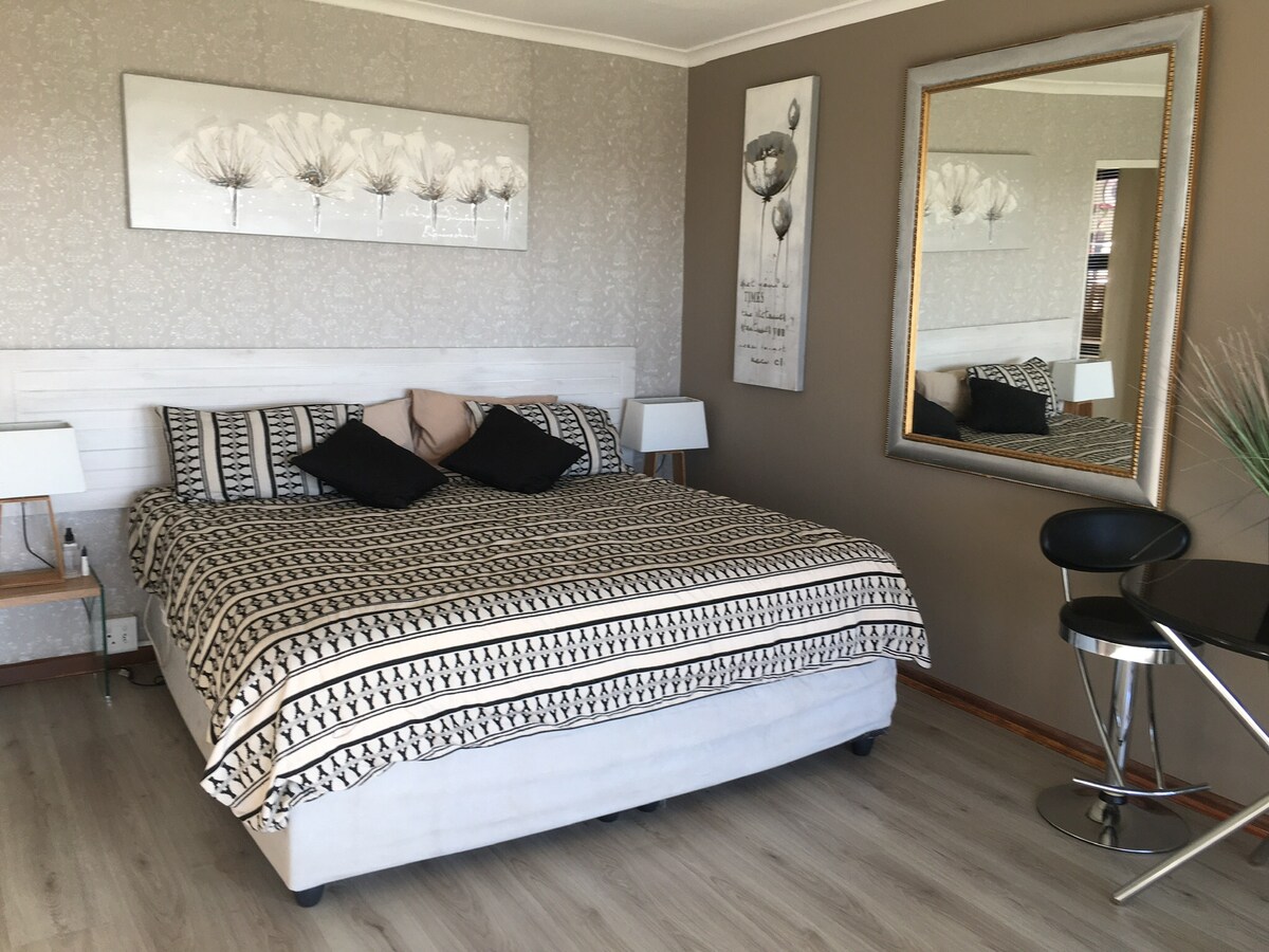 A spacious sleeping area features a large bed adorned with a patterned coverlet and several decorative pillows. A mirror reflects the room's design, which includes light-colored walls and wooden accents. A small dining table with two chairs is positioned to one side.