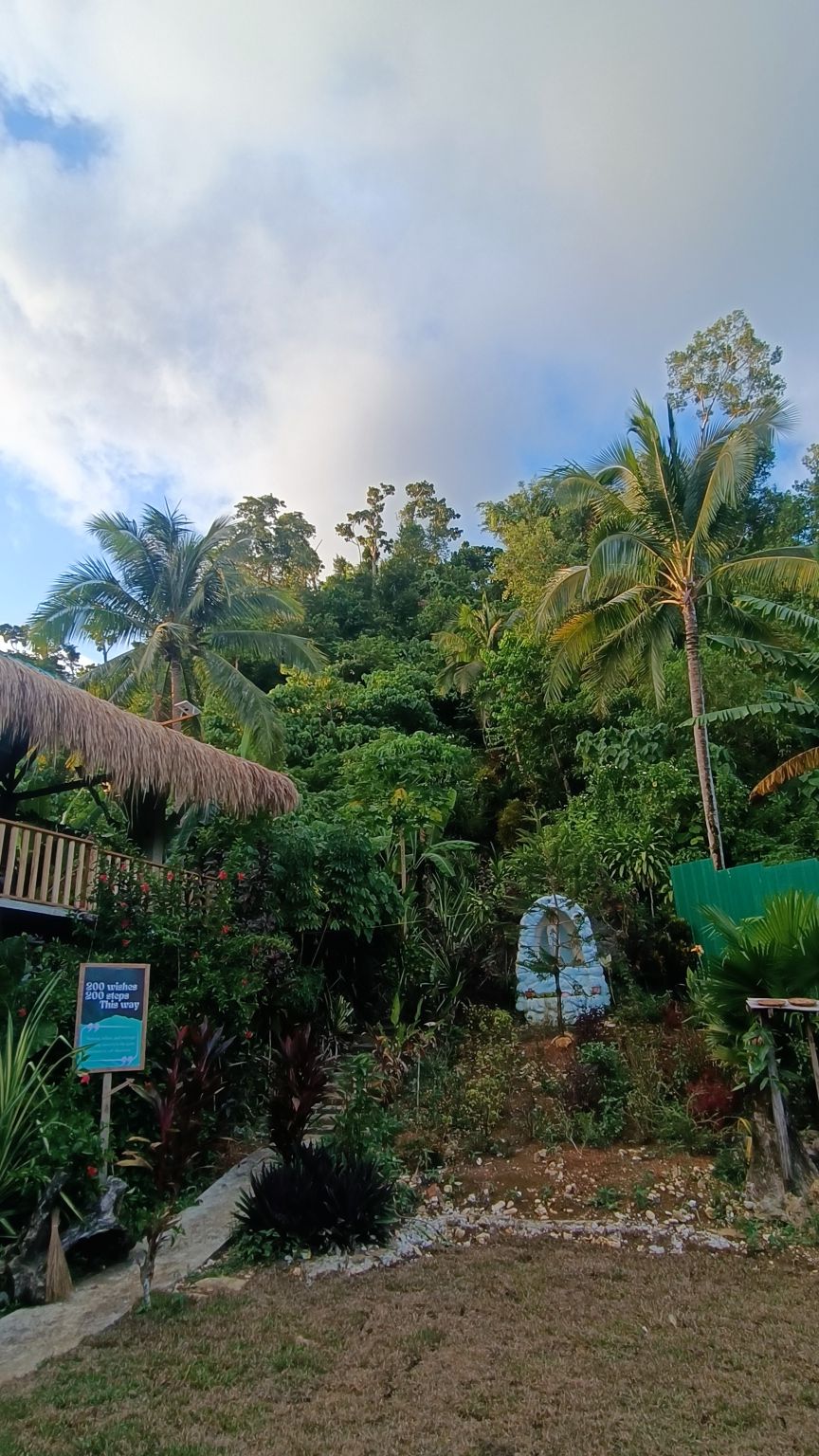 Lush tropical landscaping features diverse foliage and palm trees, creating a natural setting. A thatched-roof structure is visible on the left, with verdant greenery rising in the background. A stone marker is positioned among the plants, adding an element of interest to the serene environment.