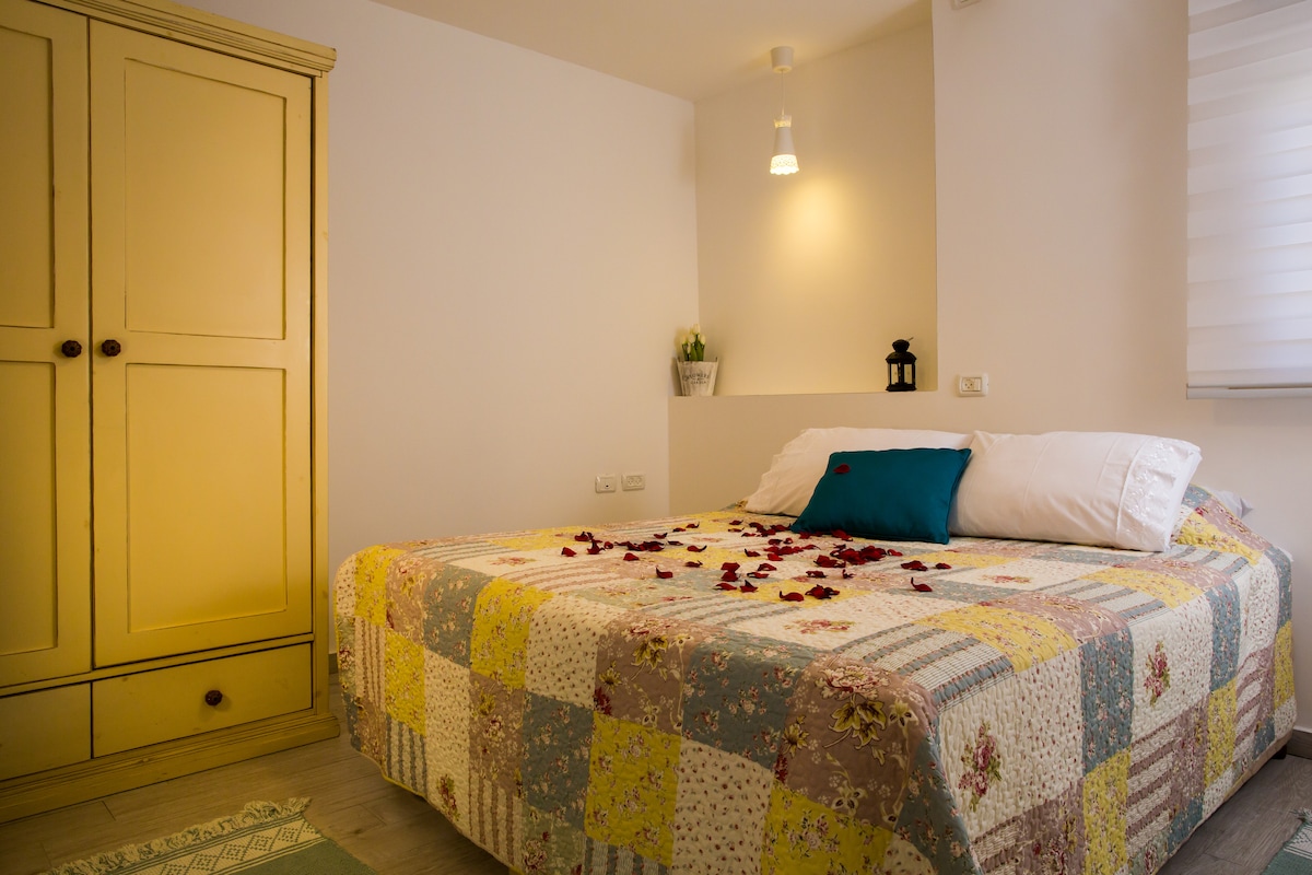 A bedroom is characterized by a vibrant, patchwork quilt covering a spacious bed. A wardrobe with a warm yellow hue stands nearby, and a soft light fixture provides gentle illumination. Rose petals are arranged artfully on the bed, adding a touch of elegance to the space.