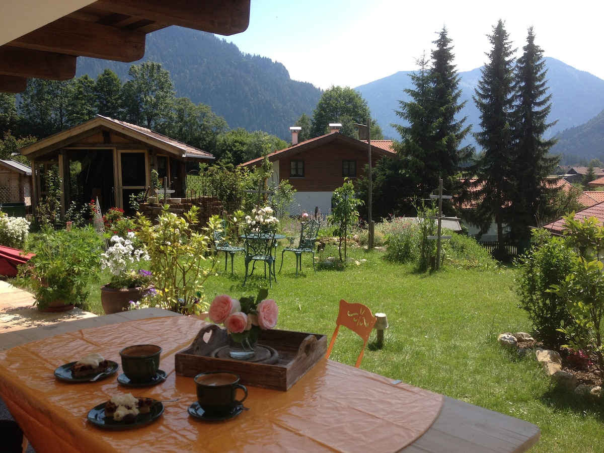 A sunny terrace features a table set with coffee cups and pastries, surrounded by a lush garden. Rolling hills and mountains are visible in the background, offering a serene view. Colorful flowers and a small wooden cabin add to the tranquil outdoor setting.