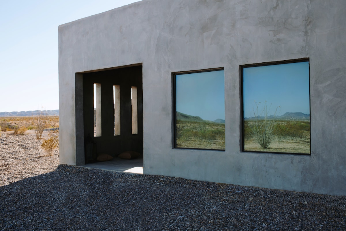 A modern casita exterior features large windows reflecting the surrounding desert landscape. The minimalist soft gray walls contrast with the earthy tones of the pebbled ground. A small entryway is visible, providing a glimpse of the inviting interior space.