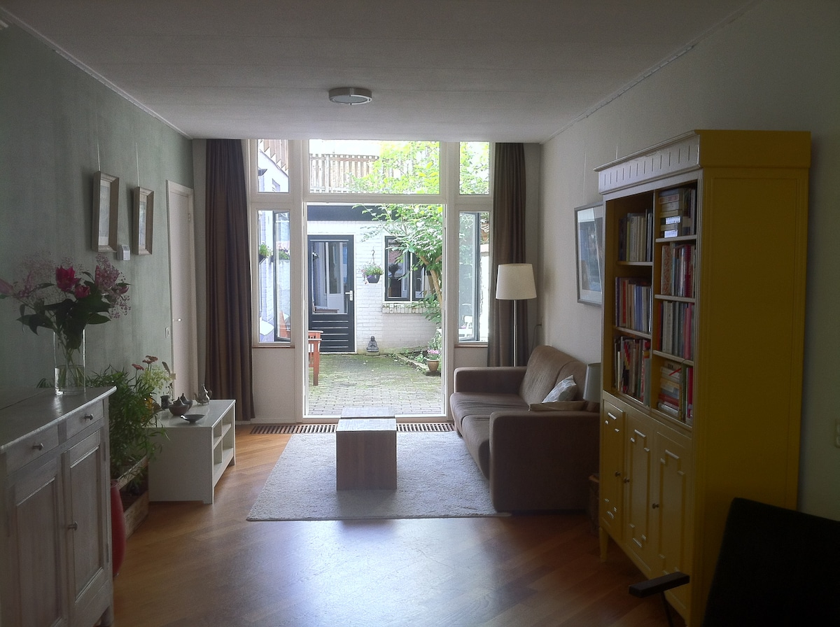 The spacious living room features a wooden parquet floor and large windows that allow natural light to fill the space. A comfortable couch and a yellow cabinet for storage are visible, along with a coffee table placed on a light rug. French doors lead to the garden area.