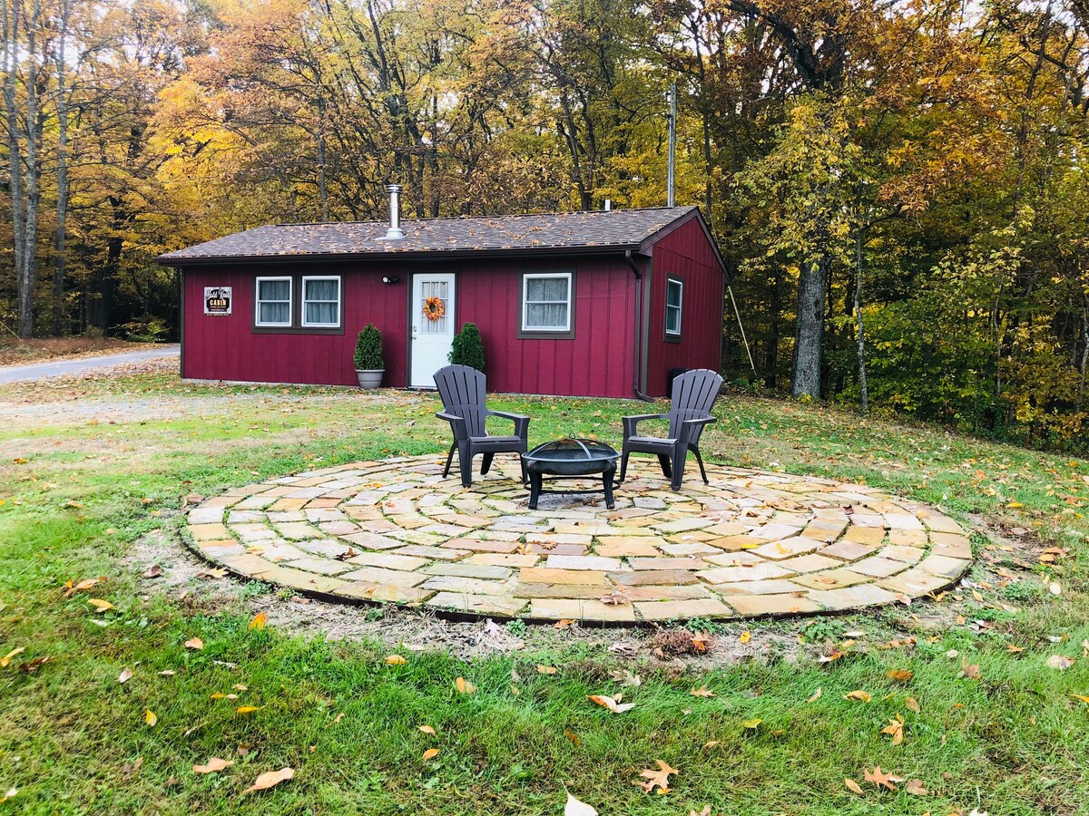 A circular patio made of stone pavers is set in front of a red cabin. Two black Adirondack chairs are positioned around a fire pit, surrounded by fallen leaves and trees with autumn foliage. The cabin features a welcoming door and flower pots by the windows.