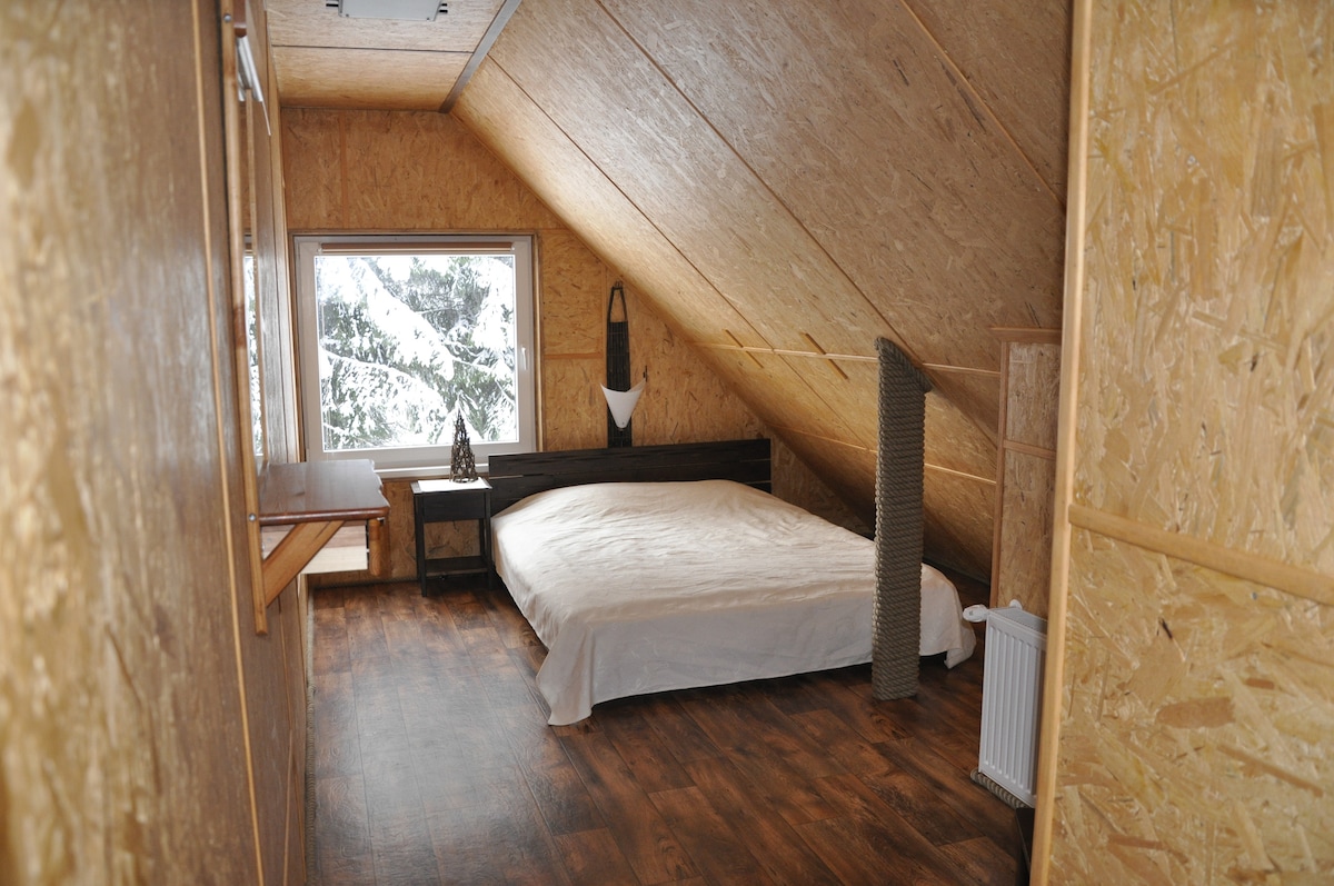A spacious room features a large bed with a light-colored blanket, positioned under a slanted ceiling. Natural light filters through a window overlooking the snowy landscape. Wooden walls and flooring create a warm atmosphere, accompanied by minimal furnishings, including a small desk and heater.