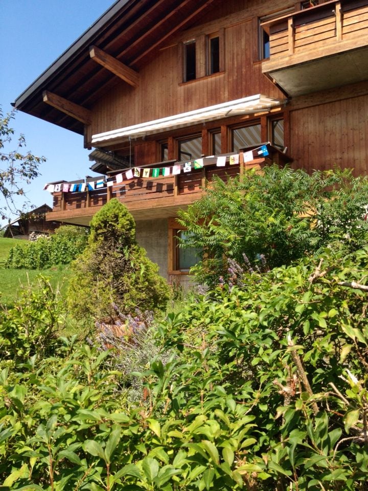 The exterior of a wooden chalet features a balcony adorned with colorful banners. Lush greenery surrounds the building, creating a natural landscape. The structure has multiple levels and showcases a blend of woodwork and concrete, under a clear blue sky.