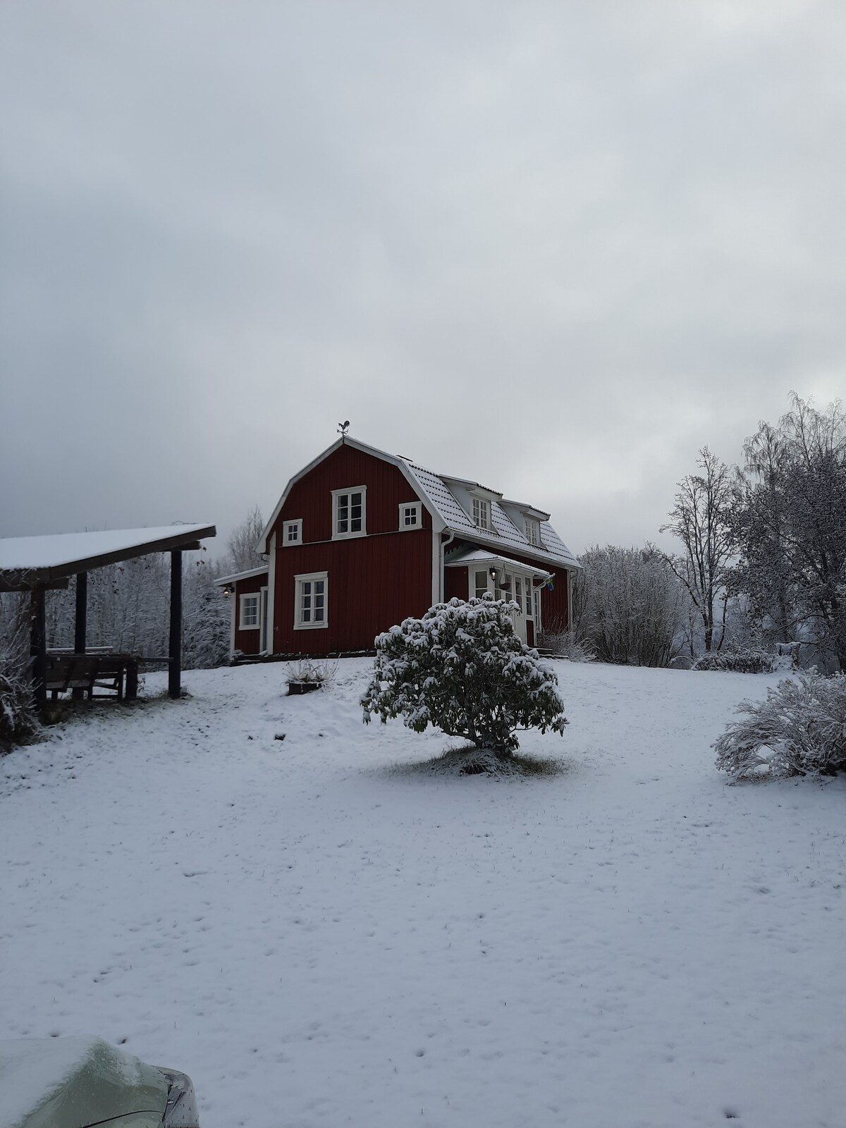 A traditional red house is seen blanketed in fresh snow, surrounded by a serene, winter landscape. The roof is dusted with white snow, and a small tree adds interest in the foreground. Trees in the background are frosted, enhancing the peaceful setting.