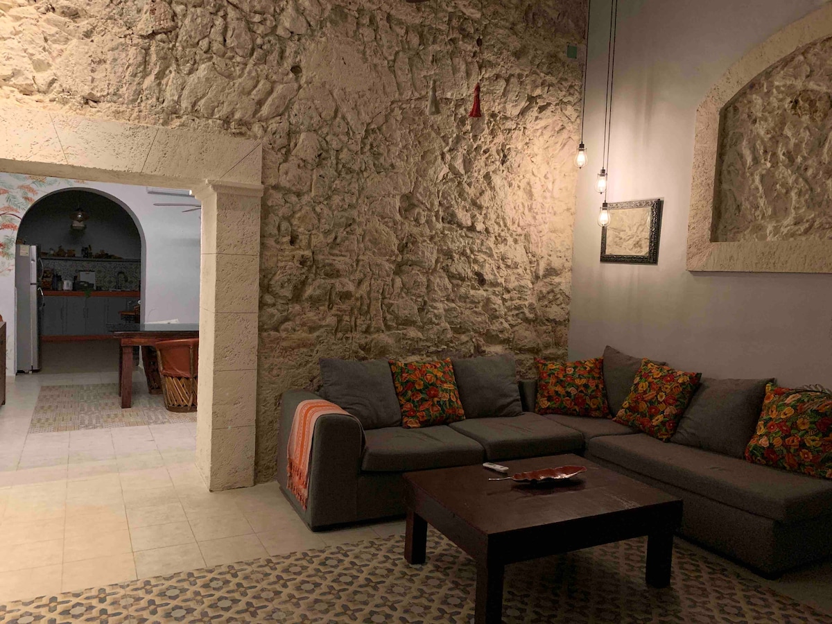A cozy living area features a comfortable sofa adorned with colorful cushions, set against a textured stone wall. A wooden coffee table is centrally positioned, while a softly lit archway leads to the kitchen and dining area in the background.