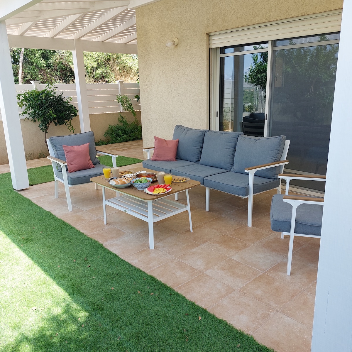 An outdoor seating area features a light-colored sofa with coordinating armchairs and decorative cushions. A wooden coffee table showcases an array of snacks and drinks. The space is surrounded by greenery, with ample natural light filtering through nearby windows.