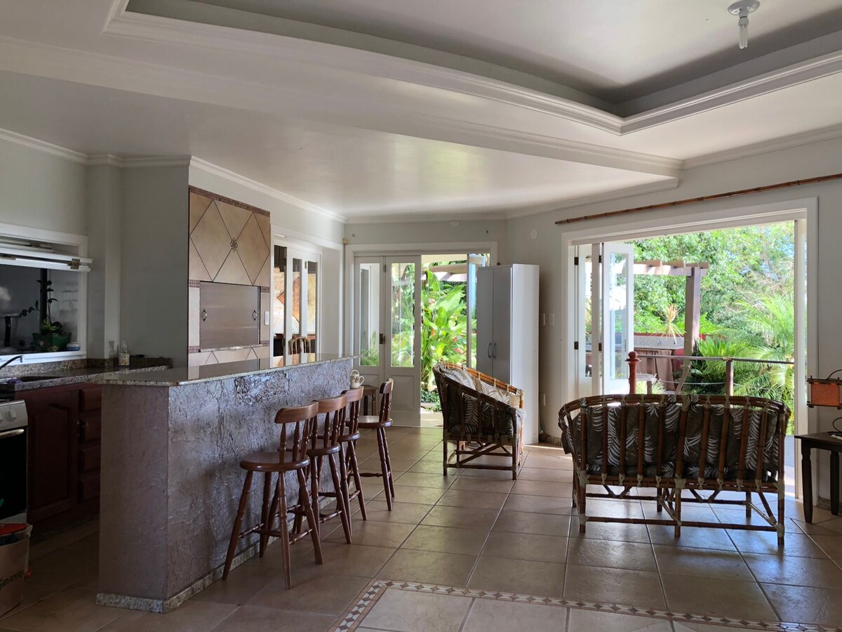 The open living area features a wide bar with seating for several guests. Natural light fills the space through large glass doors that lead to the garden. Comfortable seating arrangements are positioned around the room, offering views of the surrounding greenery.