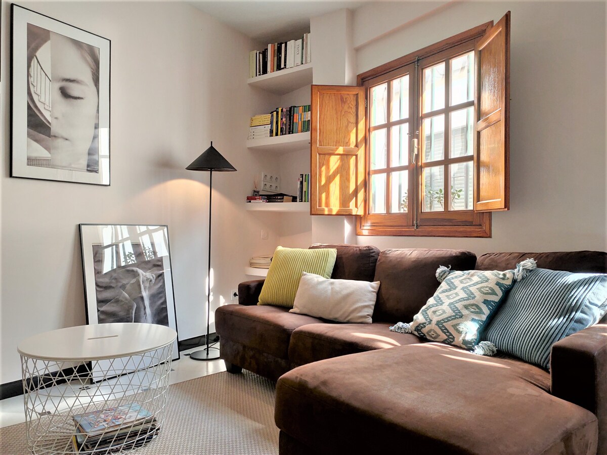 A cozy living area features a spacious brown sofa adorned with assorted cushions. Natural light filters through a wooden window, illuminating a small round coffee table and a tall lamp. Books are displayed on a nearby shelf, enhancing the inviting atmosphere.