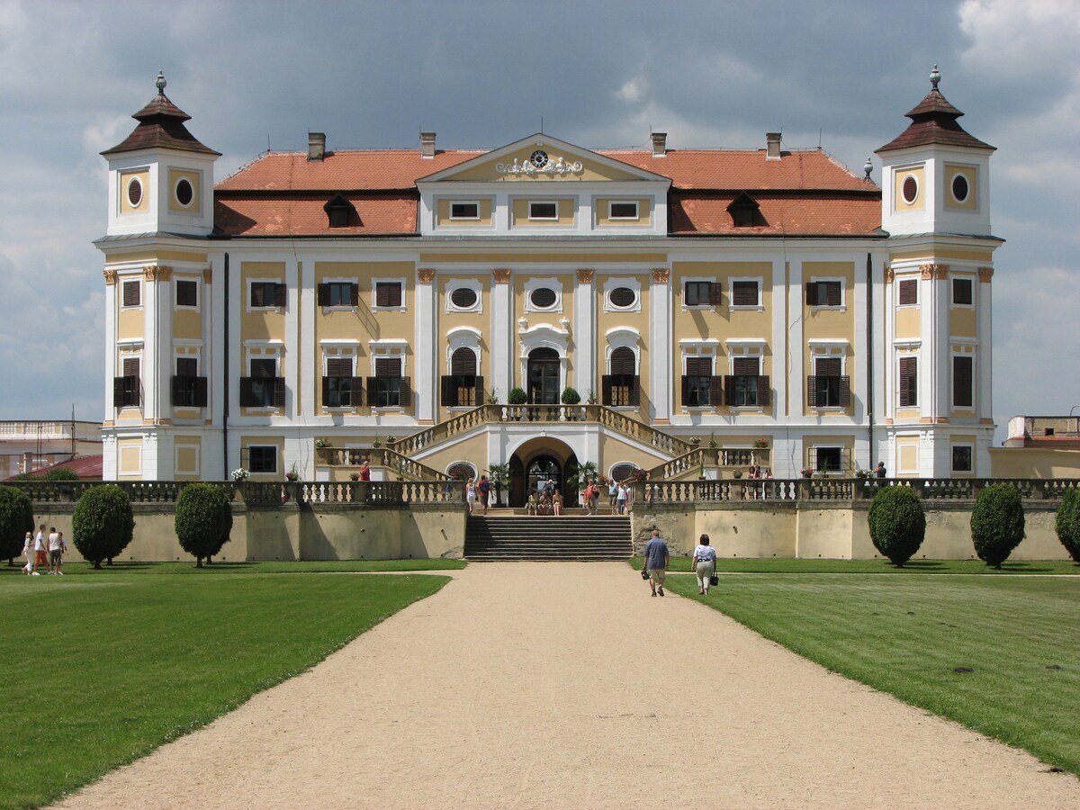 An elegant historical building is set against a cloudy sky. The baroque-style structure features ornate details, prominent towers, and an expansive staircase leading to the main entrance. Lush greenery surrounds the property, while a gravel pathway guides visitors towards the entrance.