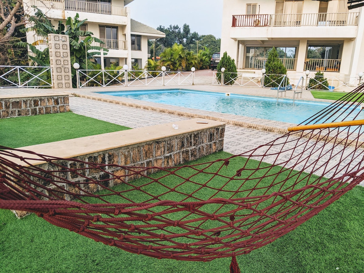 A hammock is positioned near a spacious outdoor swimming pool, surrounded by well-maintained grass. The pool area features a stone border and is flanked by lounge chairs. Lush greenery and the villa's balconies are visible in the background.