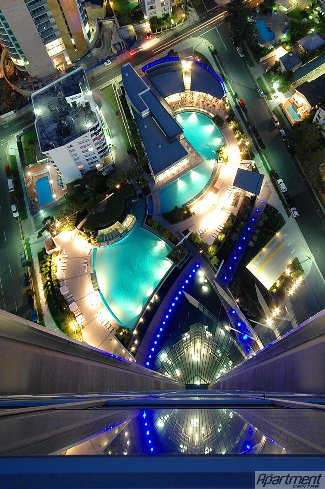 An aerial view showcases the illuminated pools and surrounding architecture, with vibrant blue lighting highlighting the outdoor amenities. The scene captures the layout of the resort area, with streets and buildings visible in the background, contributing to the urban atmosphere.