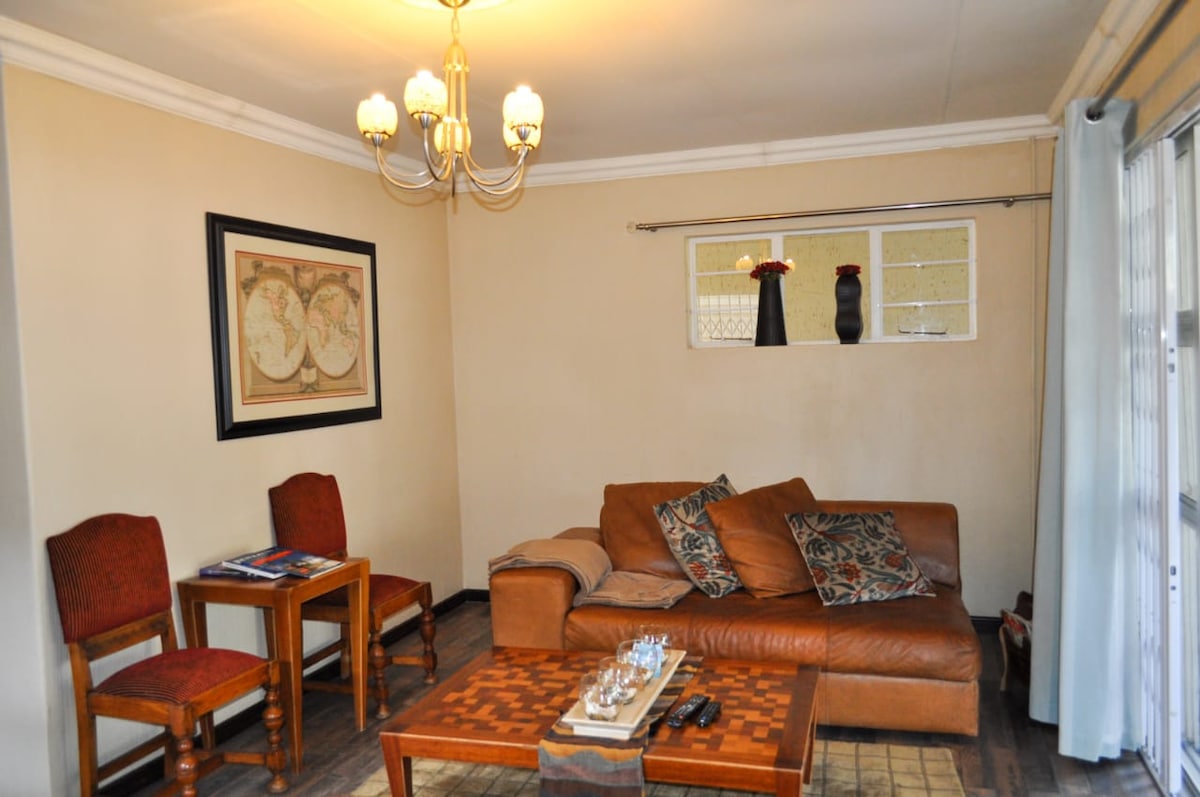 A cozy living area features a comfortable brown sofa adorned with decorative cushions. Two wooden chairs are positioned beside a framed wall art piece. A coffee table sits in front, with a decorative tray placed atop it. Natural light is filtered through the window, accentuated by light curtains.