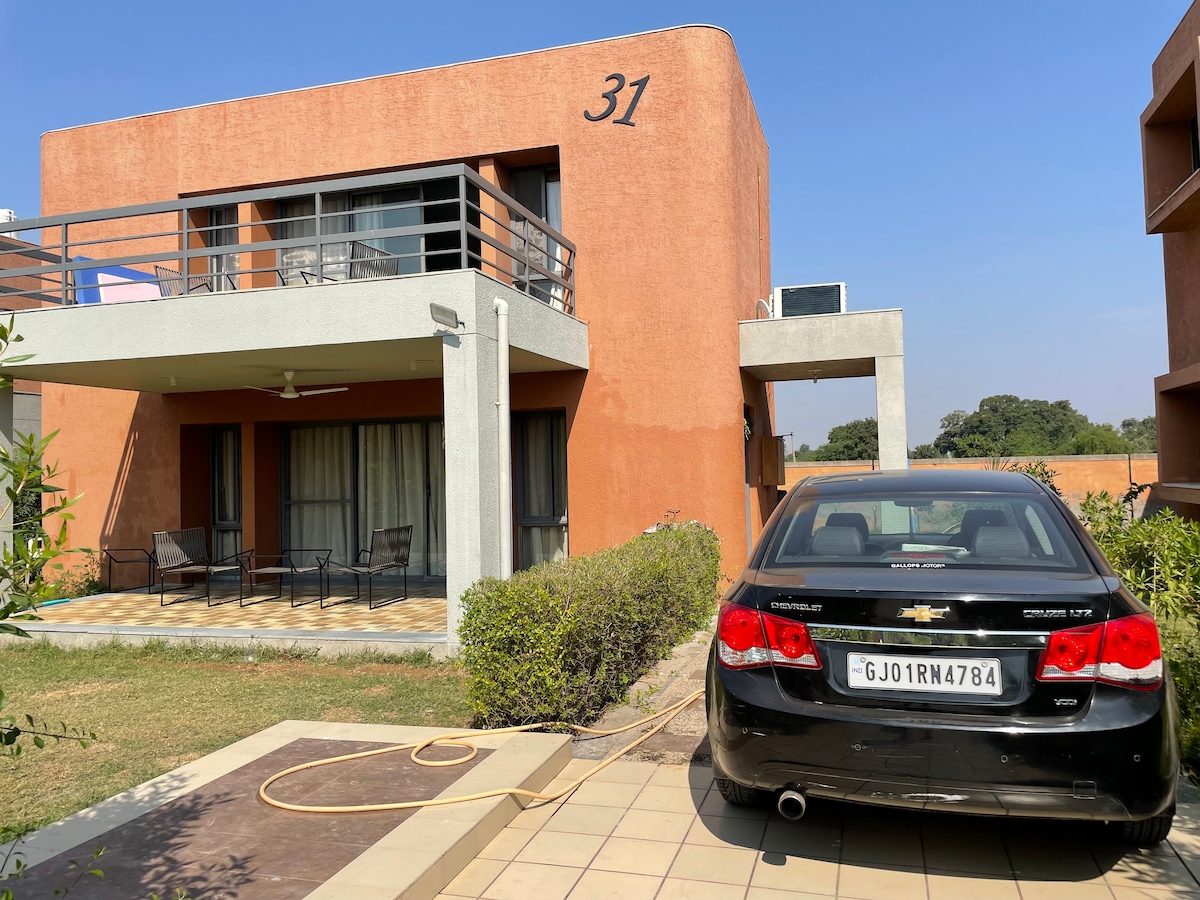 A two-story villa is featured, with a terracotta exterior and large windows. A black car is parked in front of the property, which is surrounded by well-maintained greenery. An outdoor patio area is visible, furnished with seating and a ceiling fan overhead.