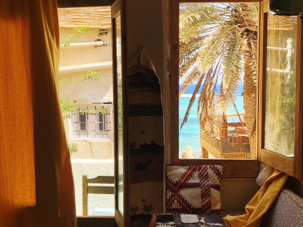 A bright and airy space features large windows with golden drapes, offering a view of the sea through palm branches. Handcrafted cushions adorn a cozy seating area, creating an inviting spot for relaxation. Sunlight filters through, illuminating the natural textures within the apartment.