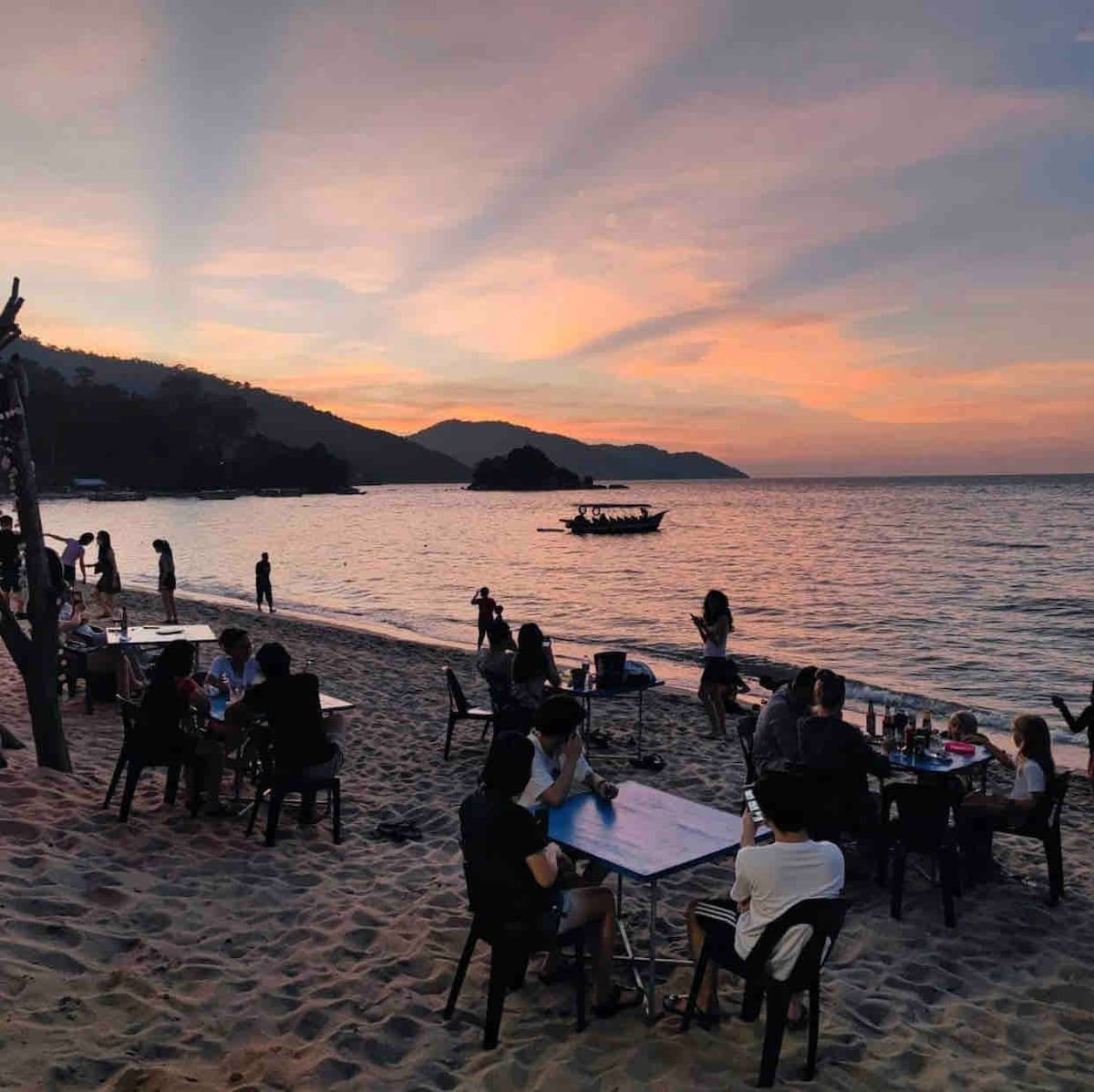The image captures people dining at tables on the sandy beach during sunset. Soft light reflects on the water, while silhouettes of guests and a boat can be seen in the distance. Gentle waves lapping the shore enhance the peaceful atmosphere.