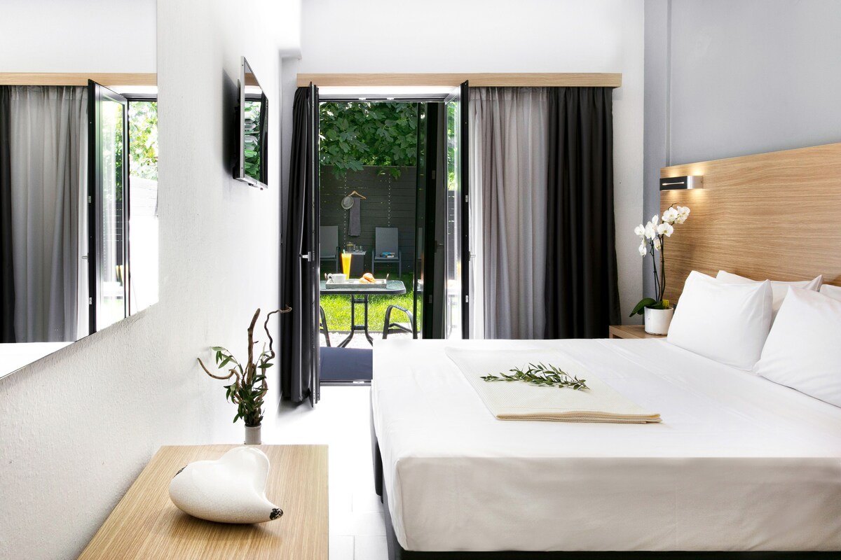 A spacious room features a large double bed with crisp white linens. Floor-to-ceiling windows open to a balcony, providing views of the garden. A small table and chairs are visible on the balcony, and a flat-screen TV is mounted on the wall.