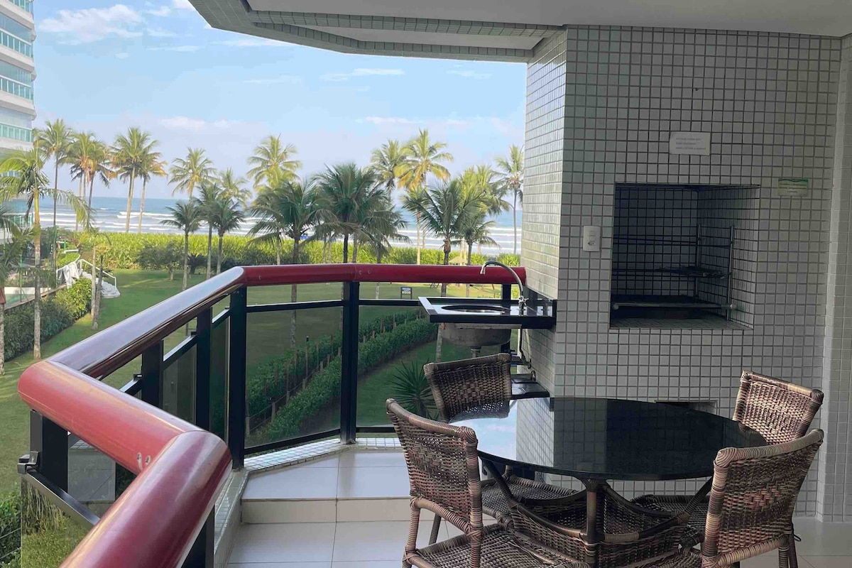 A balcony area is visible, featuring a charcoal grill and a round dining table with four wicker chairs. The glass railing offers an unobstructed view of the beach and palm trees, enhancing the connection to the serene outdoor environment.