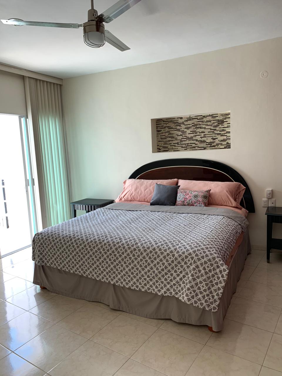 The spacious bedroom features a large bed adorned with neutral bedding and decorative pillows. Natural light floods the room through sliding glass doors that open to a small balcony, enhancing the bright and airy atmosphere. Side nightstands provide additional convenience.