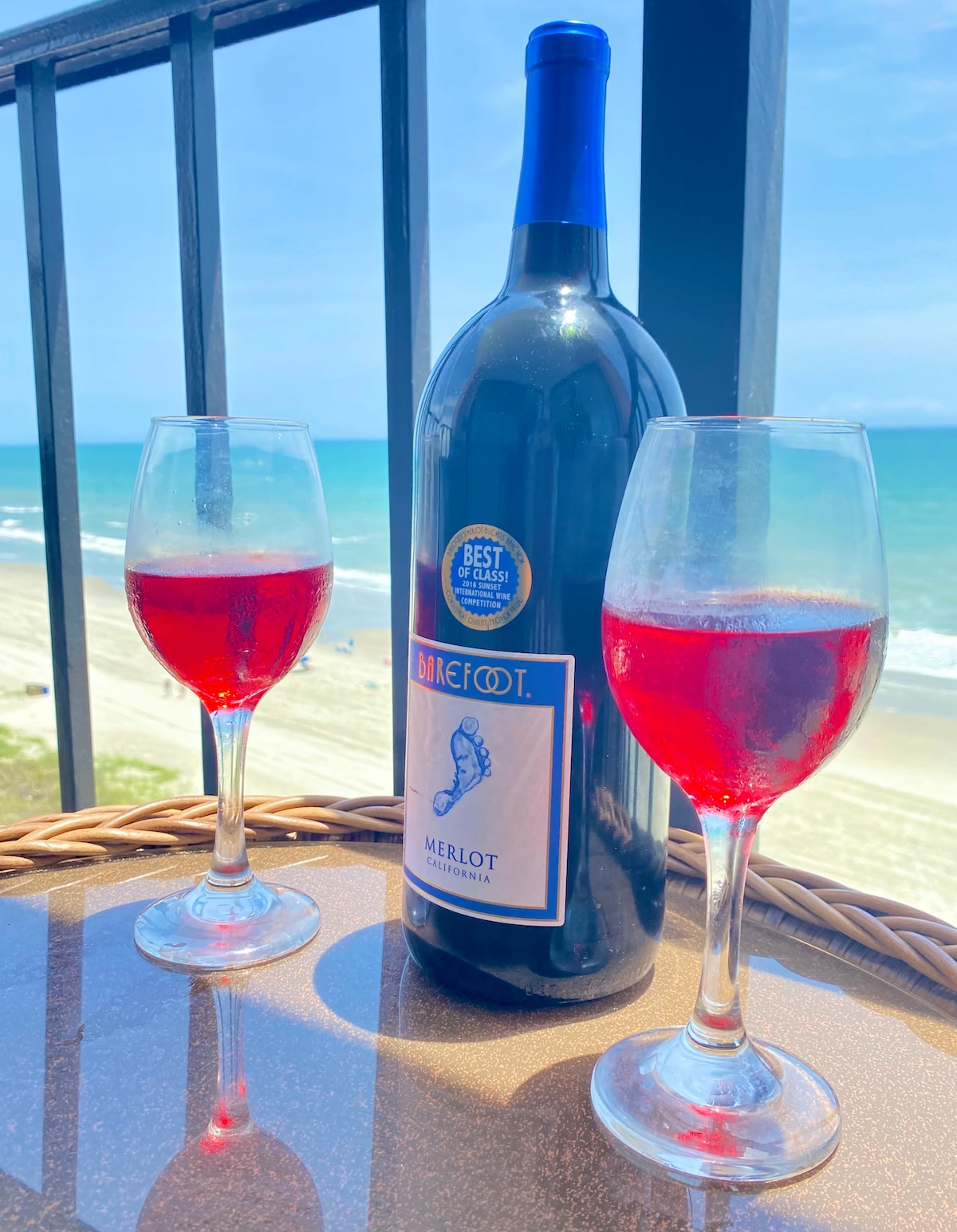 A scenic beachfront view is captured through a balcony overlooking the ocean. Two glasses filled with a red beverage sit beside a large bottle of Merlot on a glass-top table, with the sandy beach and ocean waves visible in the background.