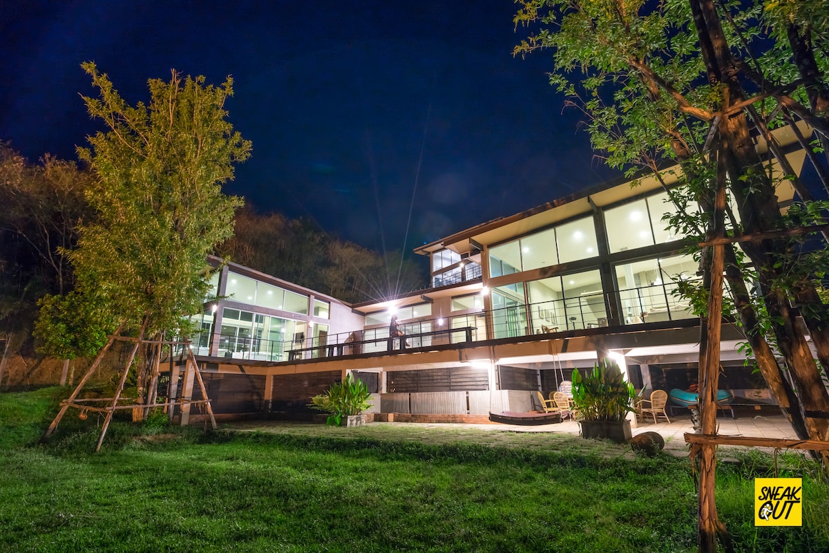 The guest house is illuminated against a night sky, showcasing its modern architecture. Large glass walls provide an open view, while a spacious patio extends outward. Surrounding greenery, including trees and plants, complements the structure, creating a natural setting.