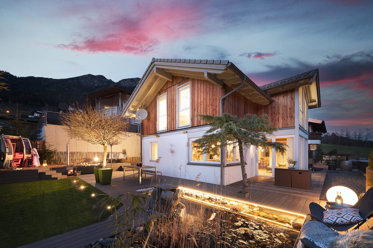 The exterior of the house features warm wood accents and large windows, blending with the natural surroundings. A tranquil garden with a pond and a deck area is illuminated by subtle outdoor lighting. The distant mountains create a scenic backdrop beneath a colorful evening sky.