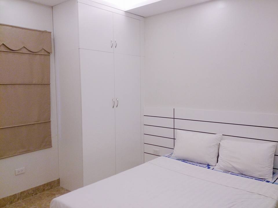 A simple and cozy room features a spacious bed with white bedding against a light-colored wall. A built-in wardrobe stands beside the bed, and a window covered with a beige curtain allows for soft natural light. The neutral tones contribute to a warm ambiance.