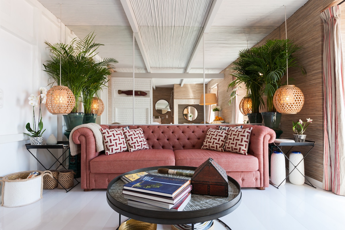 A lounge area features a Chesterfield sofa in a soft pink hue, adorned with patterned cushions. Two large potted plants flank the space, while pendant lighting provides a warm glow. A round coffee table displays books and decorative items, enhancing a relaxed atmosphere.